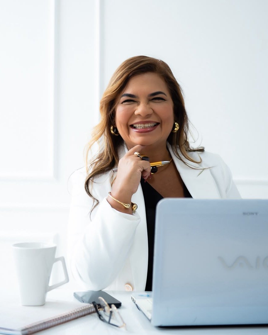 Márcia Doring sorrindo à mesa de trabalho, de blazer branco, com caneta na mão e notebook aberto