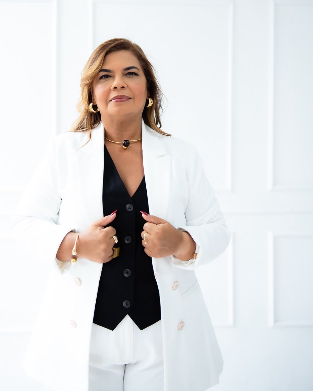 Márcia Doring em pé, de terno branco e colete preto, segurando a lapela do blazer